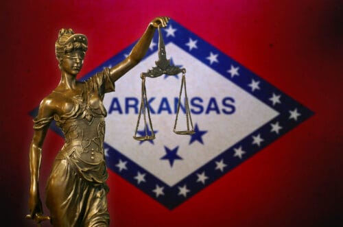 Statute of lady justice stands in front of the Arkansas flag with blue and white lozenge.
