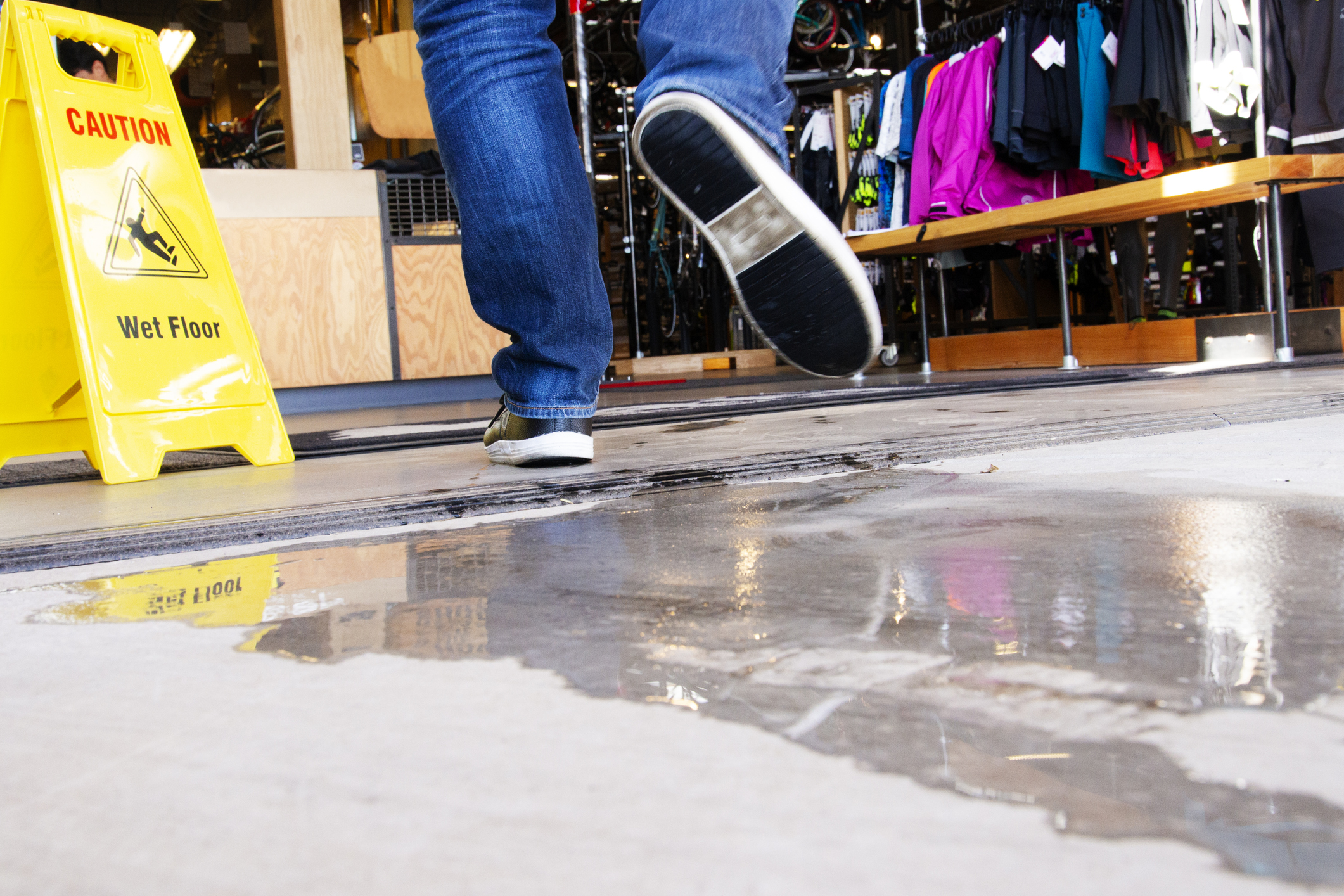 An individual is seen walking in a department store near a “Caution: Wet Floor” sign, a water puddle, and some racks of clothing.