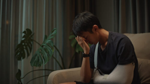 An injured man with his arm in a sling sits on the couch with his head in his hand, looking depressed.