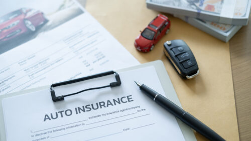 Auto insurance forms on a desk with a toy car, a fountain pen, a key fob, and stacks of cash sitting nearby.